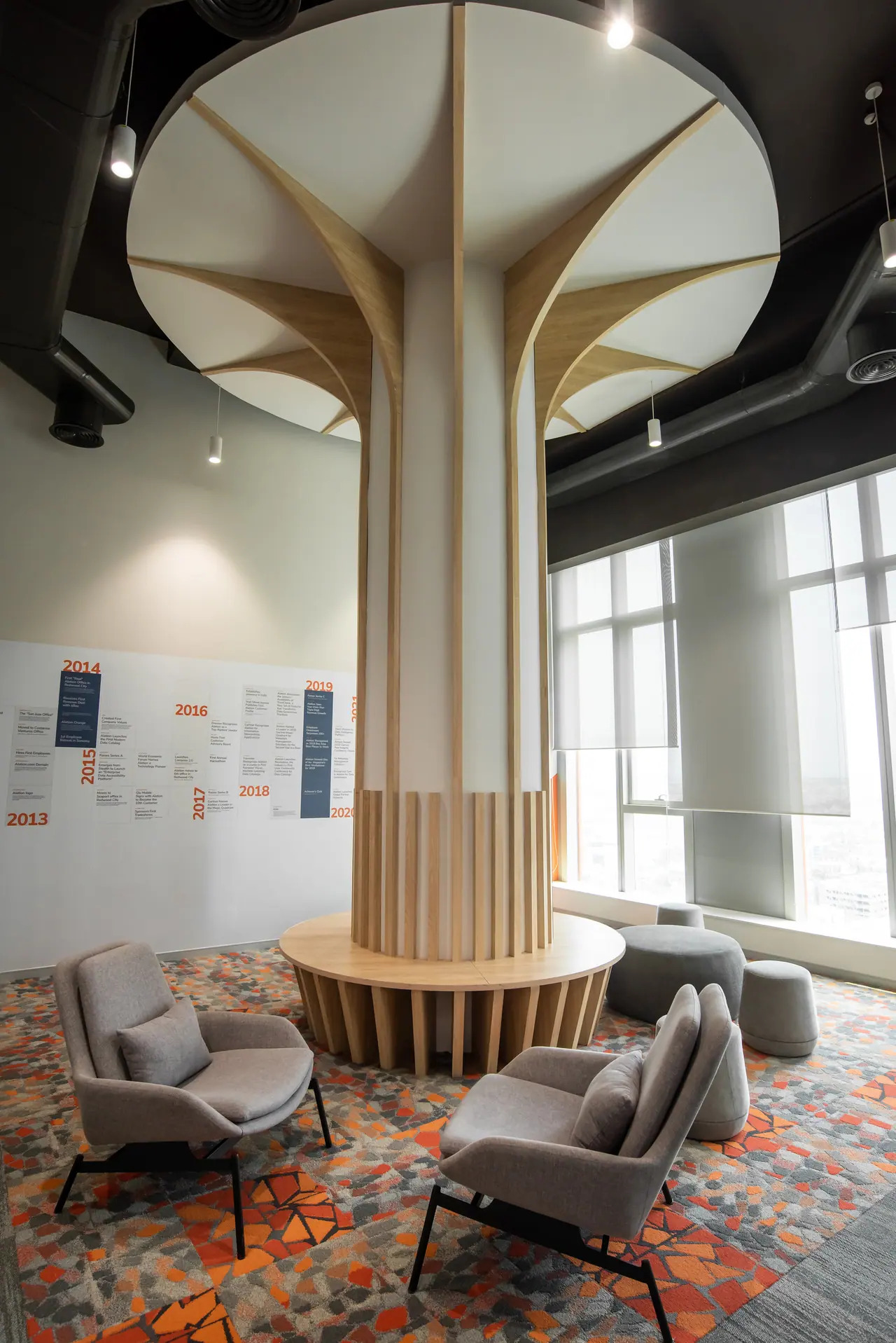Column clad as a tree with a white canopy and timber ribs over a round bench, with grey armchairs on a patterned carpet