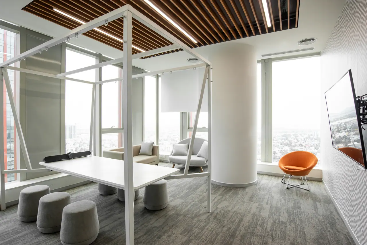 Bright collaboration room with a white-framed standing table, grey stools, sofa, orange chair and tall city-view windows