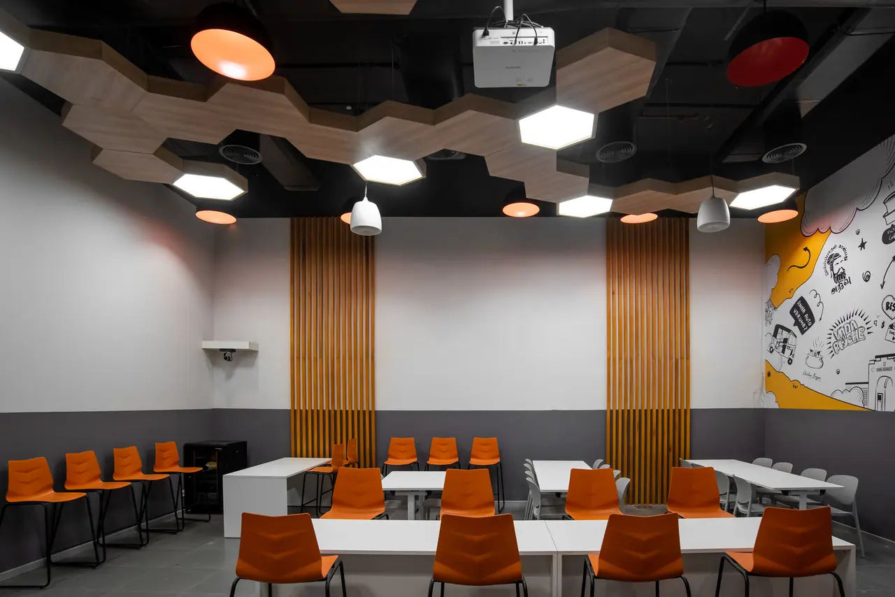 Cafeteria with orange chairs at white tables under hexagonal wood ceiling panels, timber slat strips and an illustrated mural