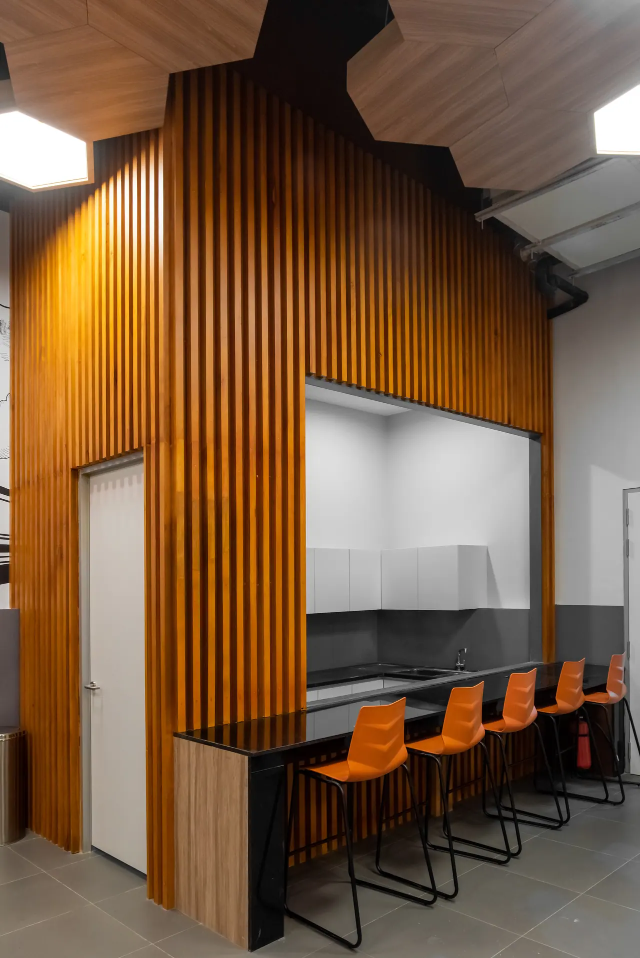 Pantry counter with orange bar stools and a black top set into a tall timber slat wall under hexagonal ceiling panels