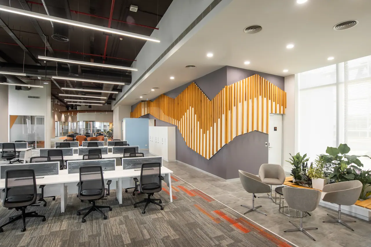 Open workstations under an exposed ceiling beside a timber batten wall cut in a mountain profile and grey lounge chairs