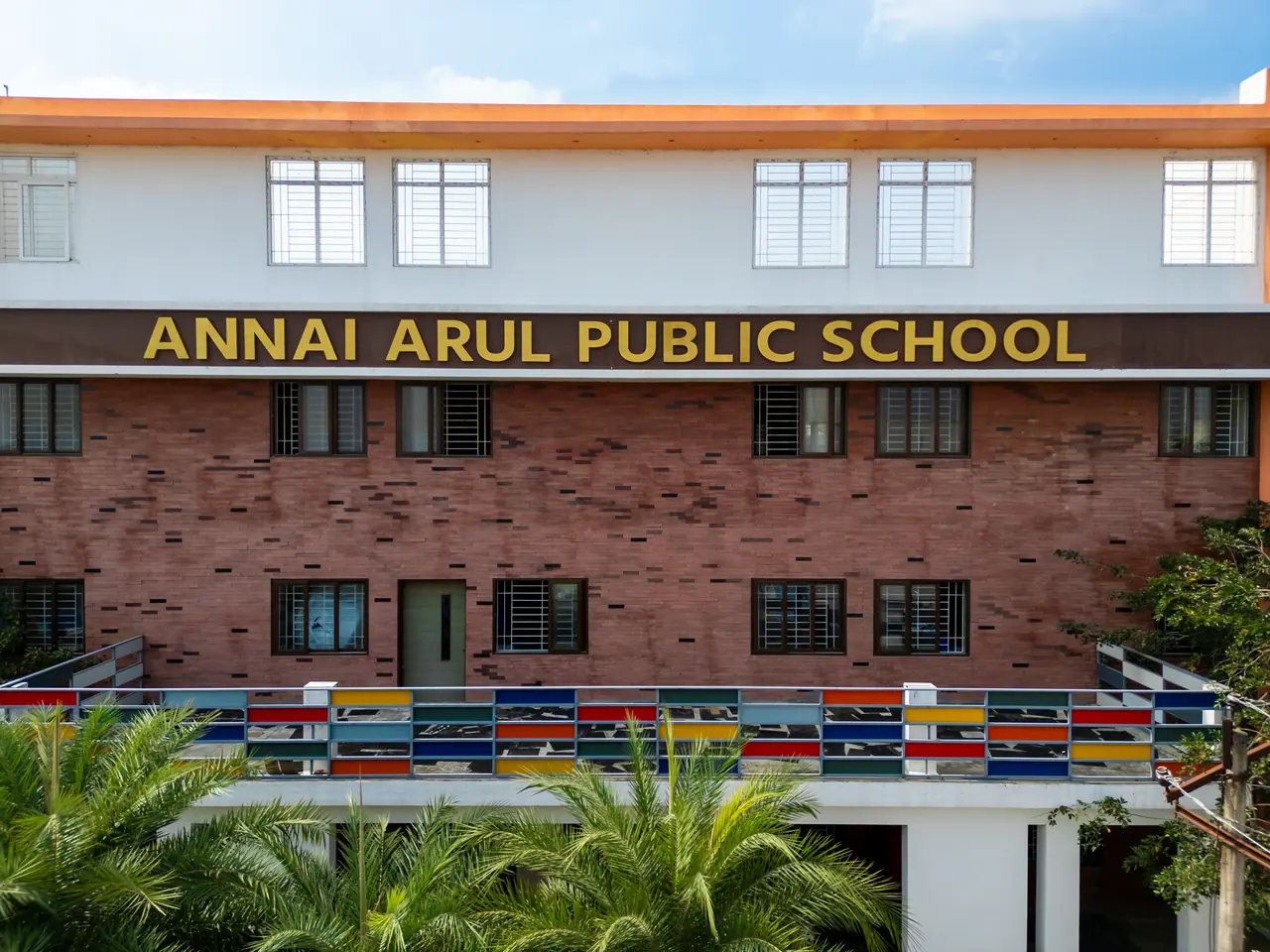 Red brick front block with gold Annai Arul Public School lettering above a balustrade of multicoloured glass panels