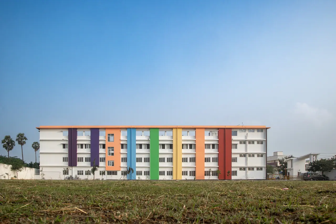 Long white school block with vertical bands of purple, orange, blue, green, yellow and red rising behind an open grass field
