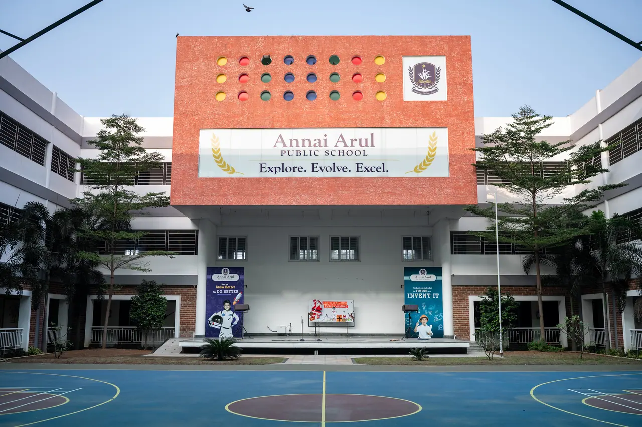 Courtyard view of a red block with colourful circular openings and school signage above a stage and a blue sports court