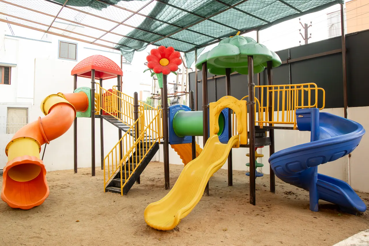 Children's playground with orange, yellow and blue slides and a flower-topped climbing frame under a green shade net