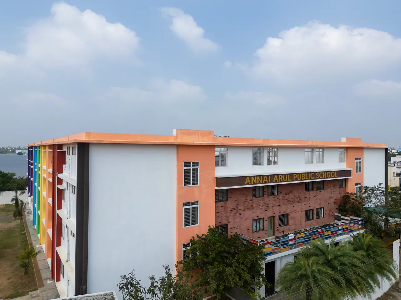 Aerial view of the school's corner: rainbow-striped side wall, red brick front block with gold lettering and palm trees