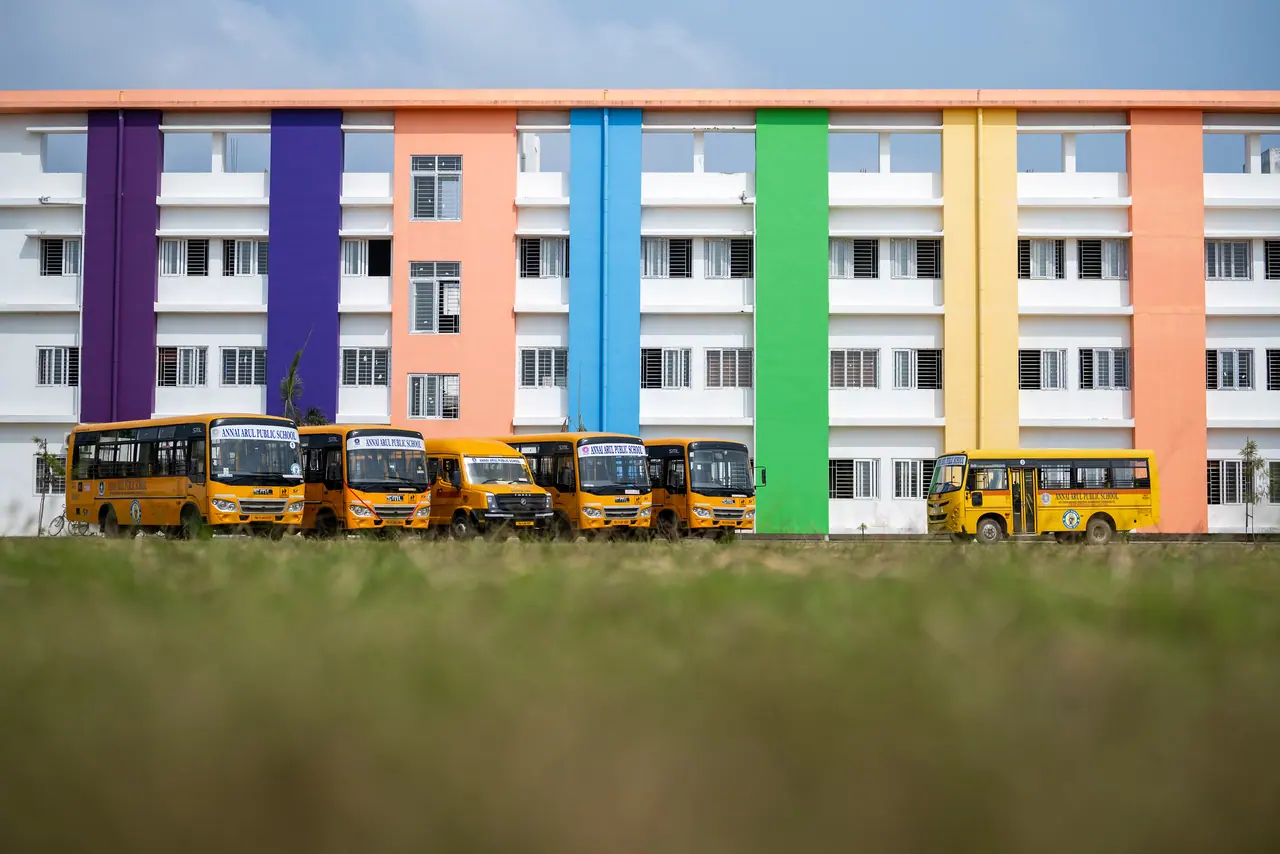 Row of yellow school buses parked in front of a white facade with vertical purple, orange, blue, green and yellow bands