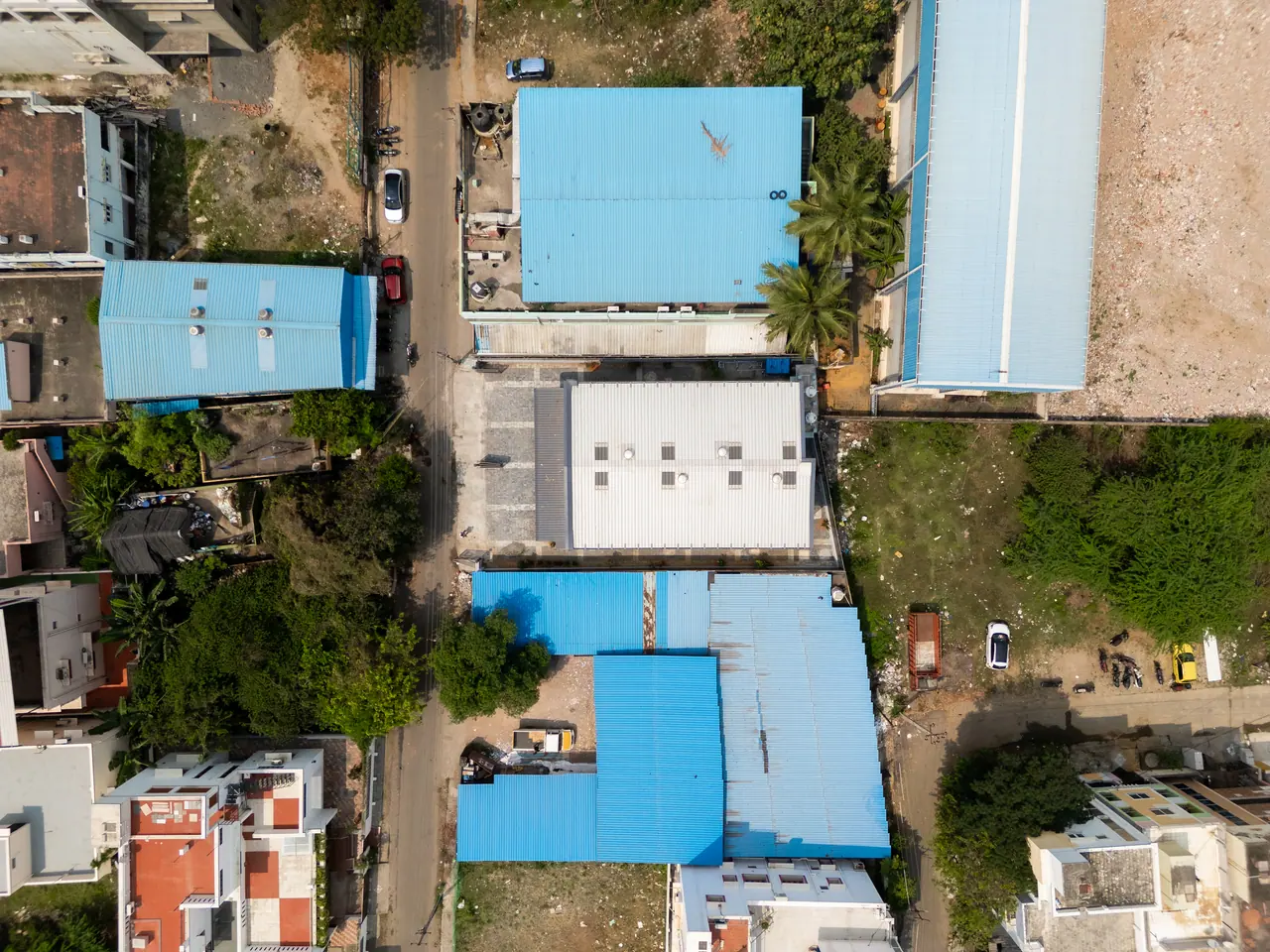 Top-down aerial view of the warehouse's pale metal roof between blue-roofed sheds, a street and open plots