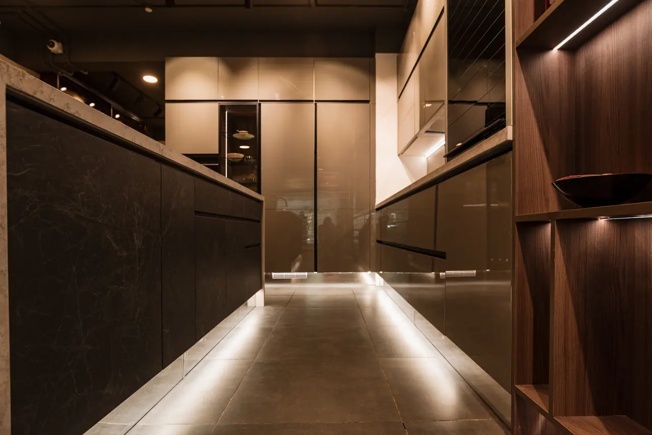 Low view along a galley between a dark marble-look island and gloss tall units, with plinth lighting on a tiled floor