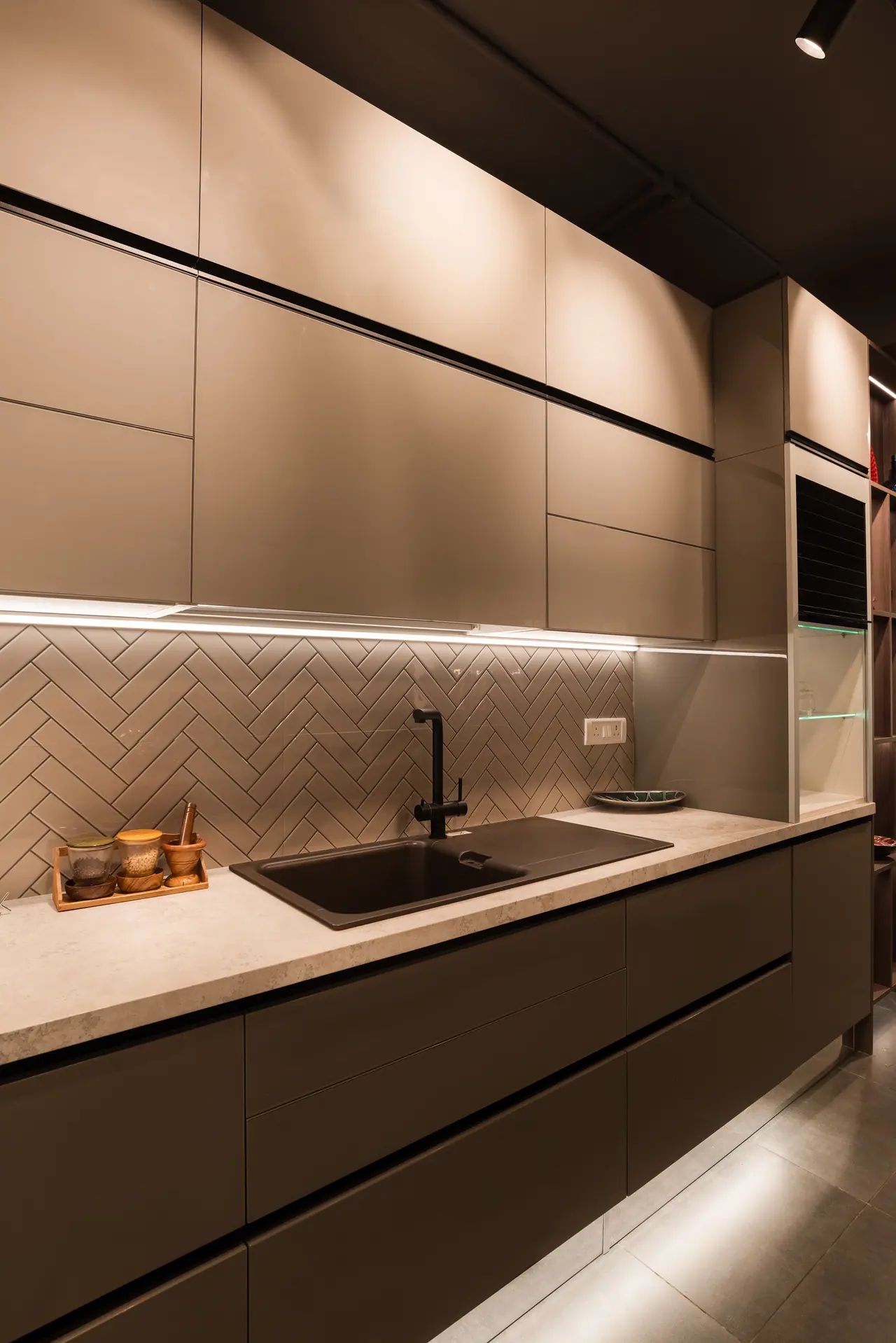 Handleless taupe kitchen run with a herringbone tile backsplash, black sink and tap, and under-cabinet strip lighting