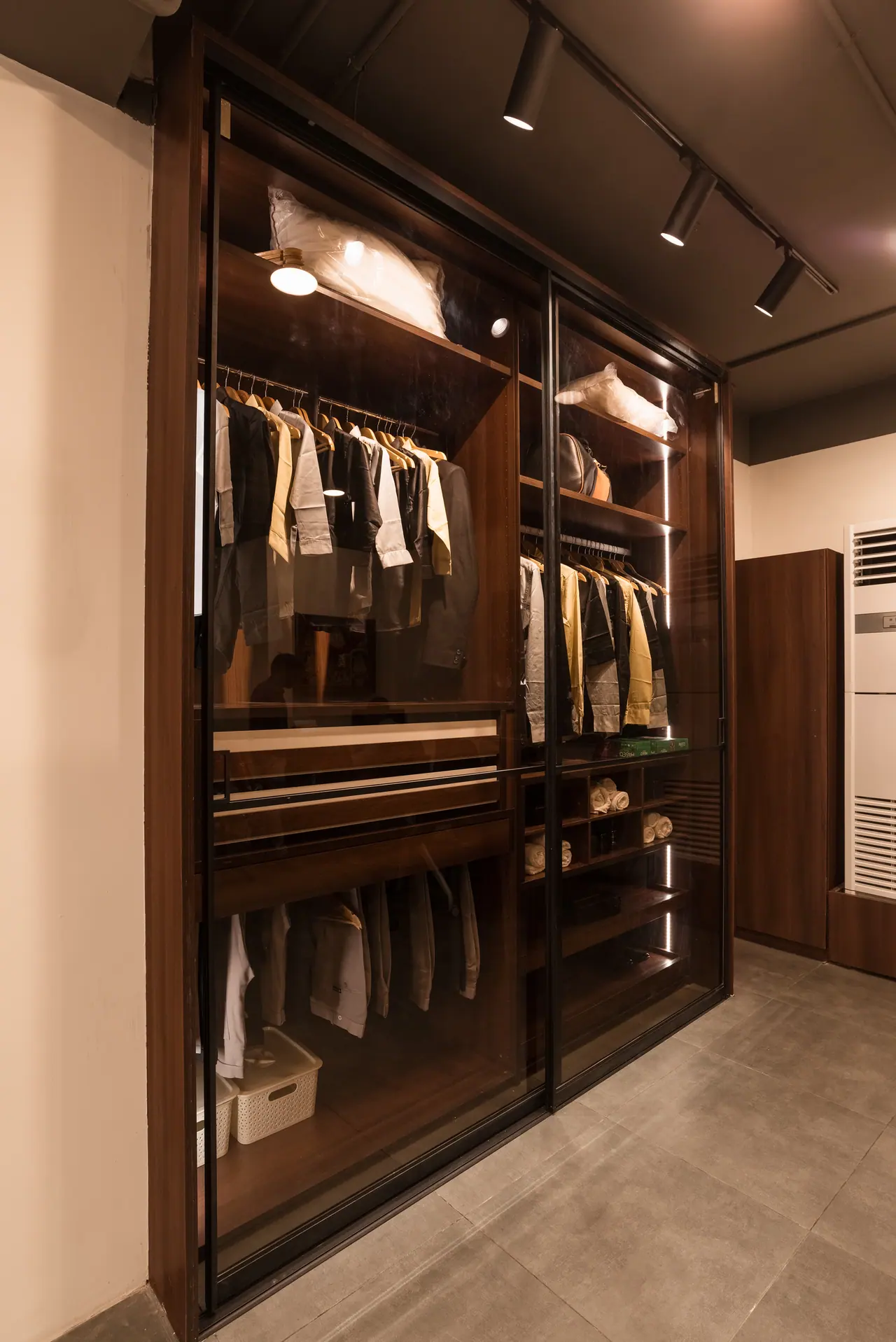 Wardrobe display with tinted glass sliding doors in a walnut frame, lit hanging rails and shelves of folded clothes