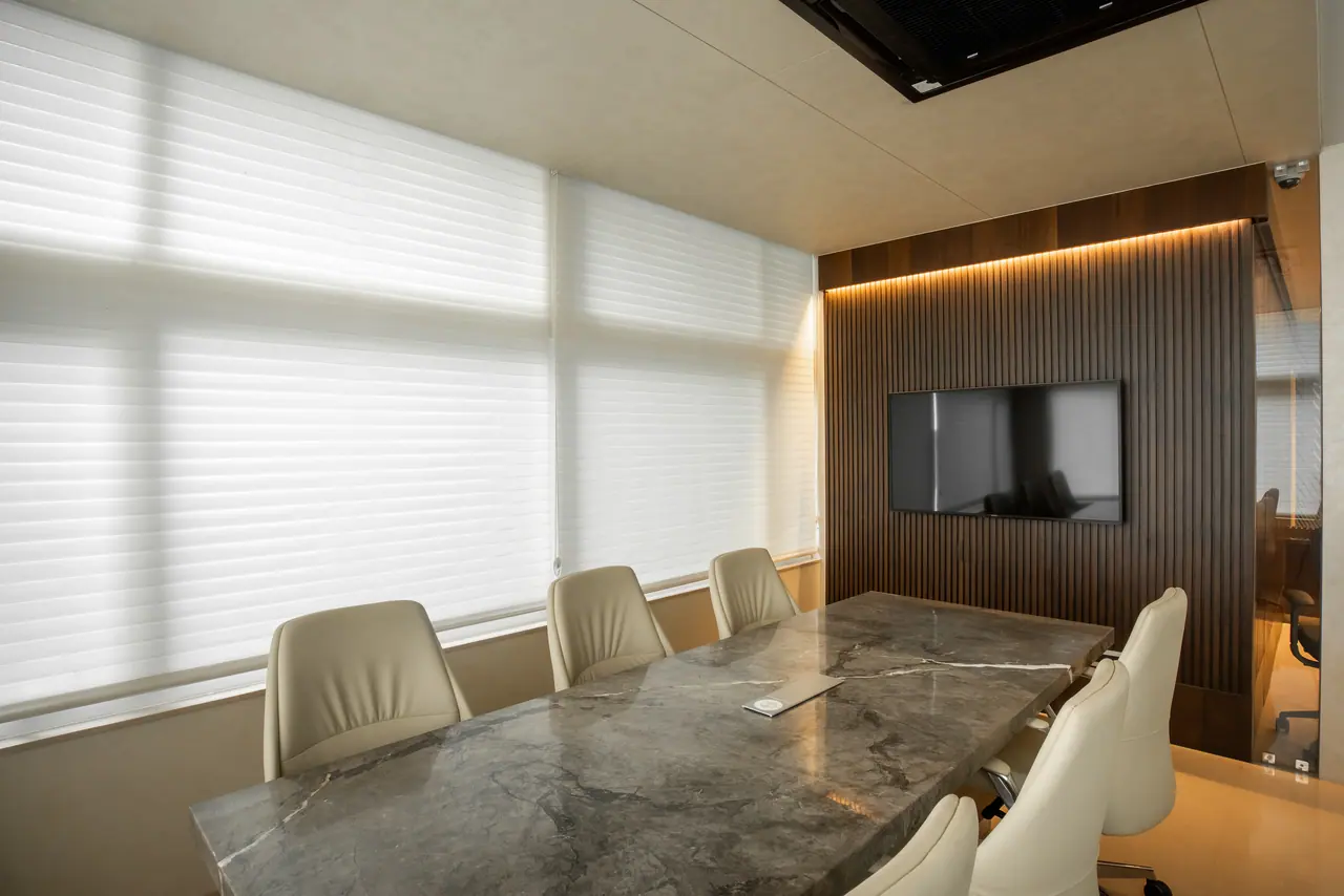 Meeting room with a grey marble table, cream leather chairs and a screen on a fluted walnut wall beside sheer blinds