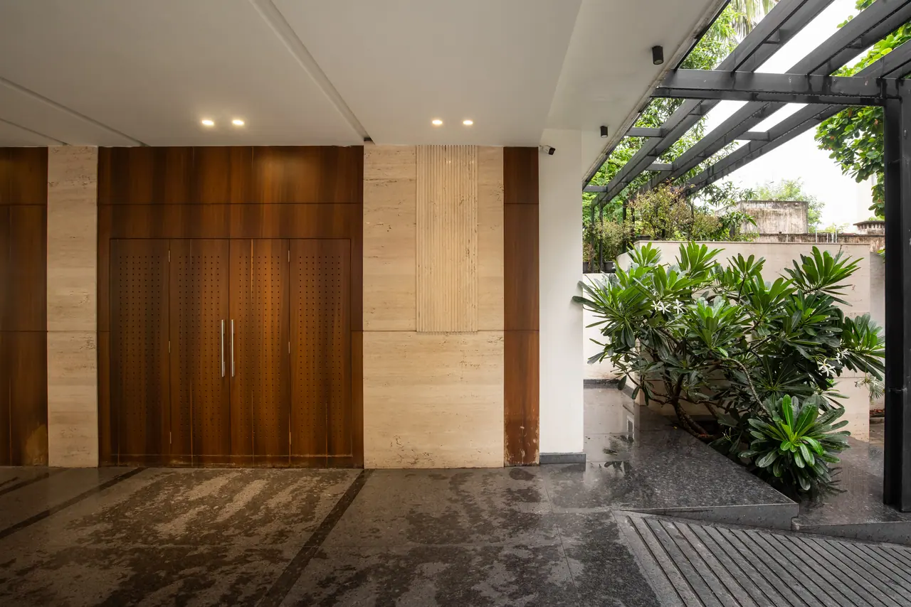 Entrance with perforated walnut double doors, travertine-look cladding, a flowering plant in a planter and a steel pergola