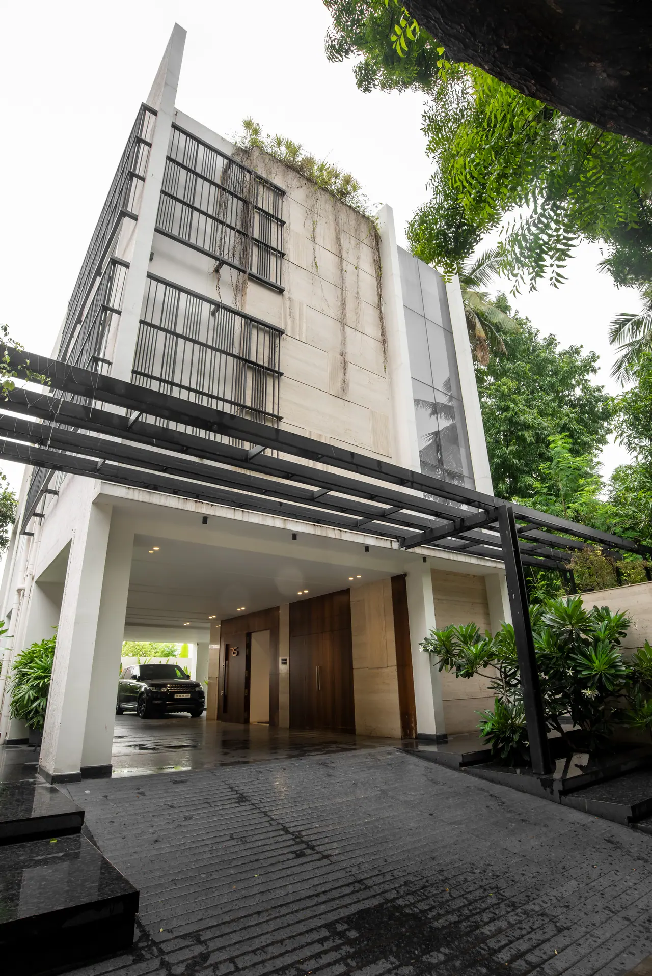 Low view of a building with a steel pergola over the driveway, stone-clad upper floors, metal grilles and trailing plants