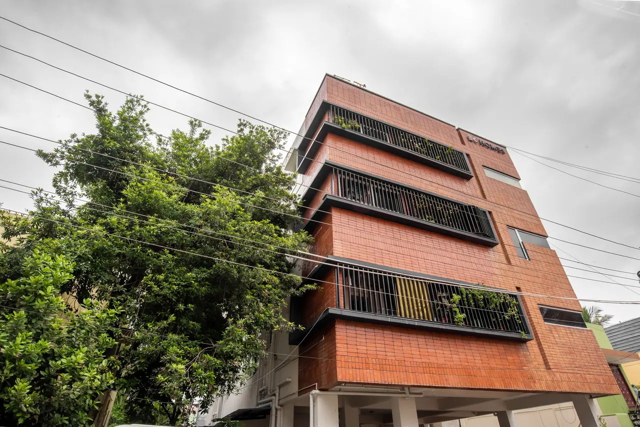 Brick-clad multi-storey building with deep black-framed balconies holding plants, seen from below beside a large tree