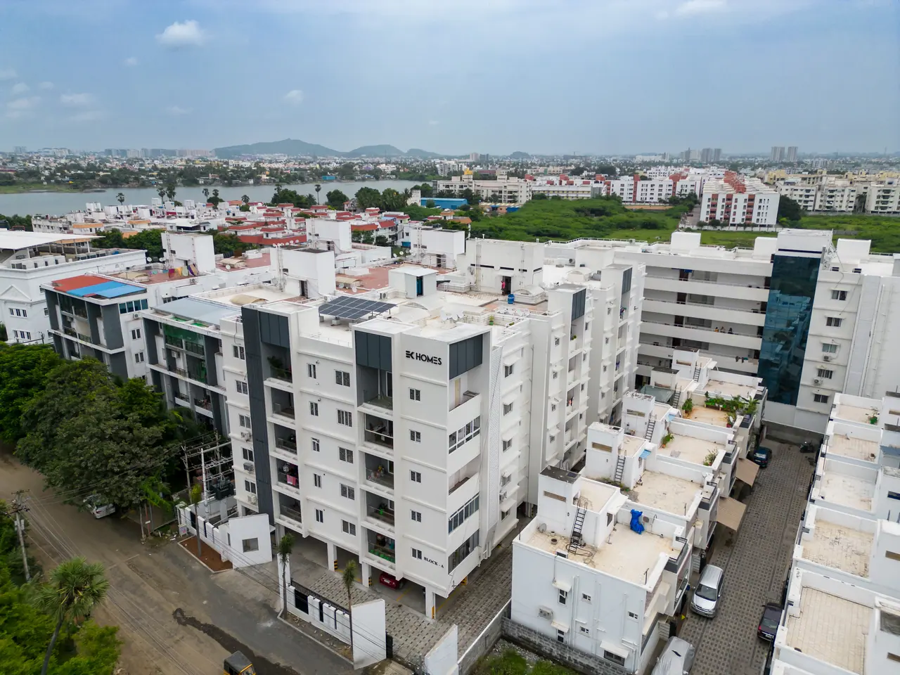 Aerial view of a white apartment block with EK Homes lettering among neighbouring buildings, with water and hills beyond
