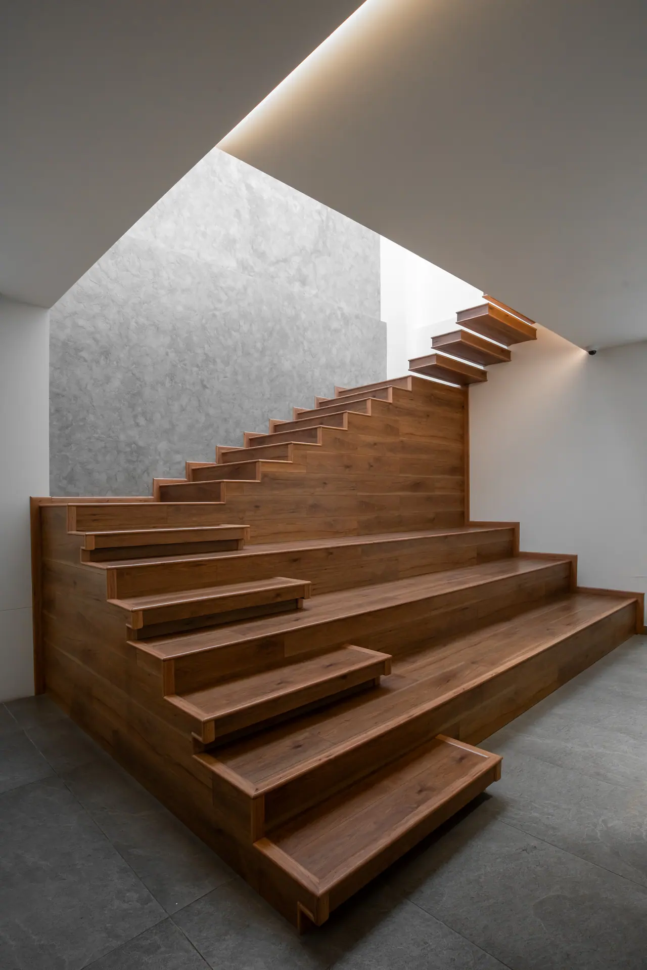 Walnut-clad staircase with cantilevered treads rising past a grey textured wall beneath a cove-lit ceiling