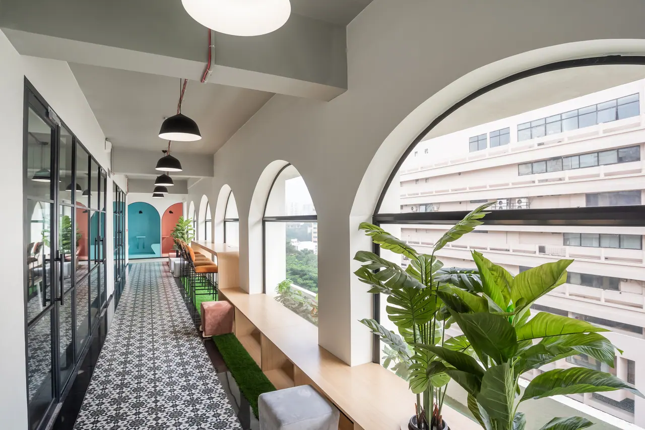 Corridor lined with arched windows and black-framed glass cabins, with patterned floor tiles, a timber ledge and plants