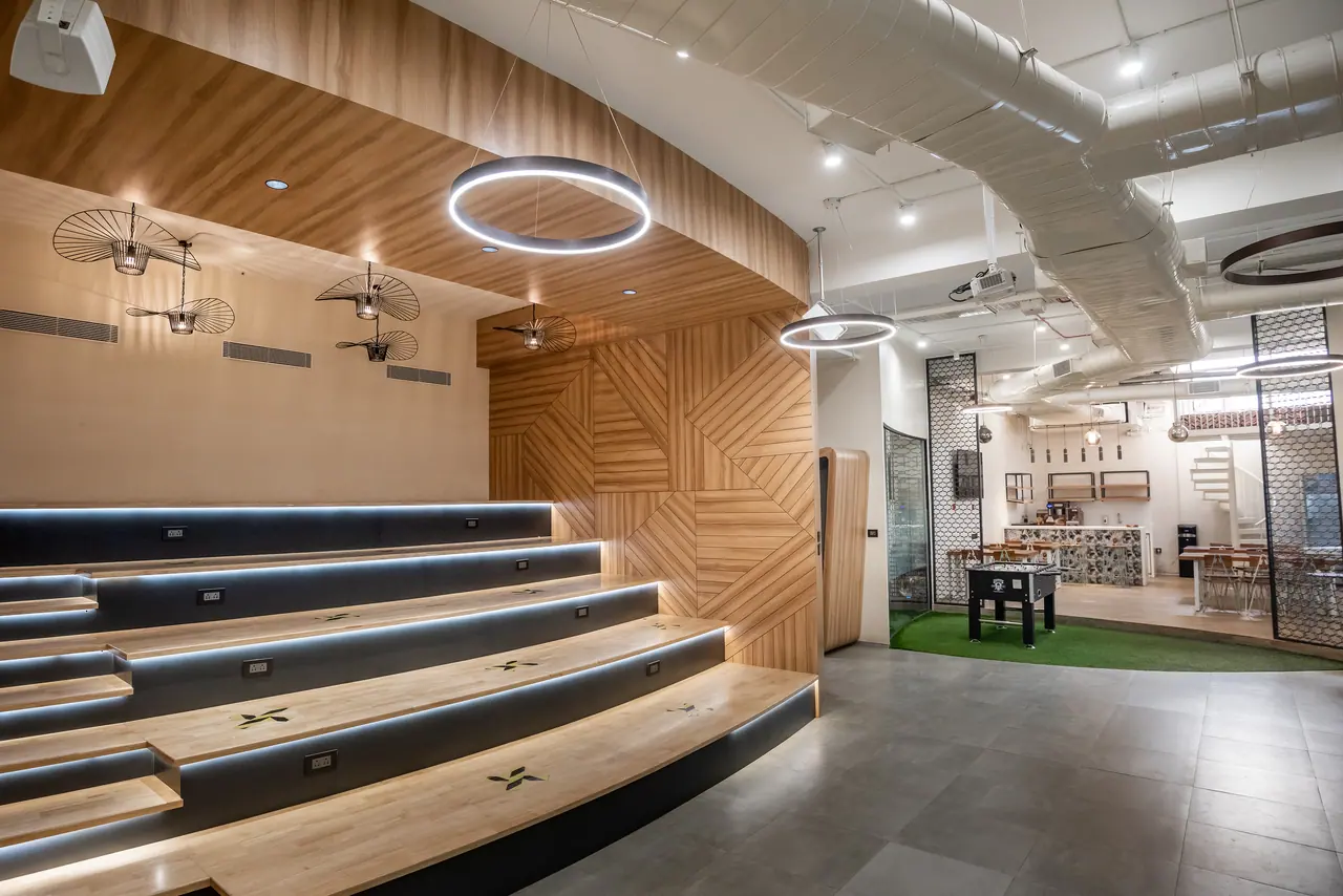 Tiered timber amphitheatre seating with lit risers under a curved wood-panelled canopy, ring pendants and a foosball table