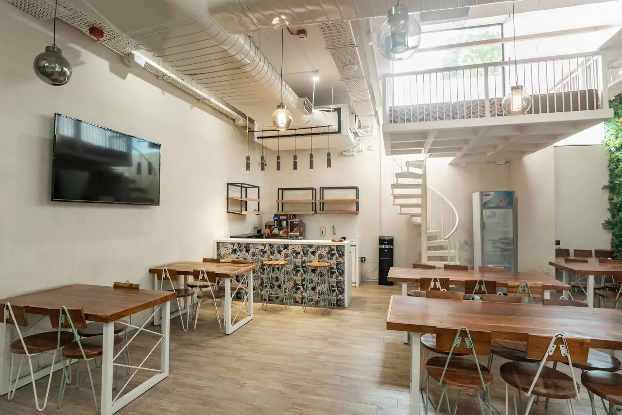 Double-height cafeteria with timber tables, a patterned-tile counter, a white spiral stair and a mezzanine with seating