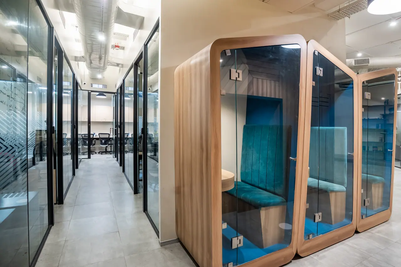 Glass-fronted phone booths with timber frames and teal upholstered benches beside a corridor of glass-walled cabins
