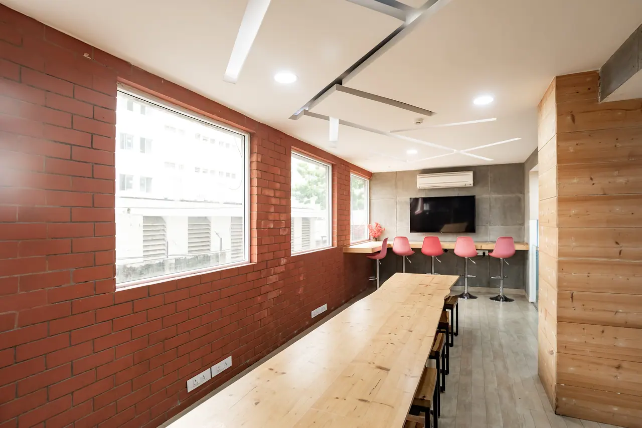 Pantry with an exposed red brick wall, large windows, a long timber table and pink bar stools at a counter under a TV