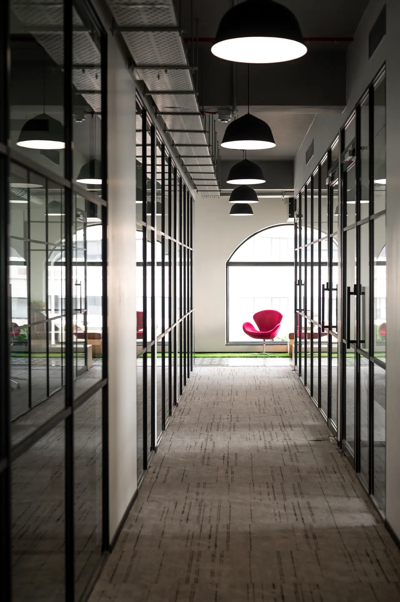 Carpeted corridor between black-framed glass cabins, with dome pendants leading to an arched window and a red chair