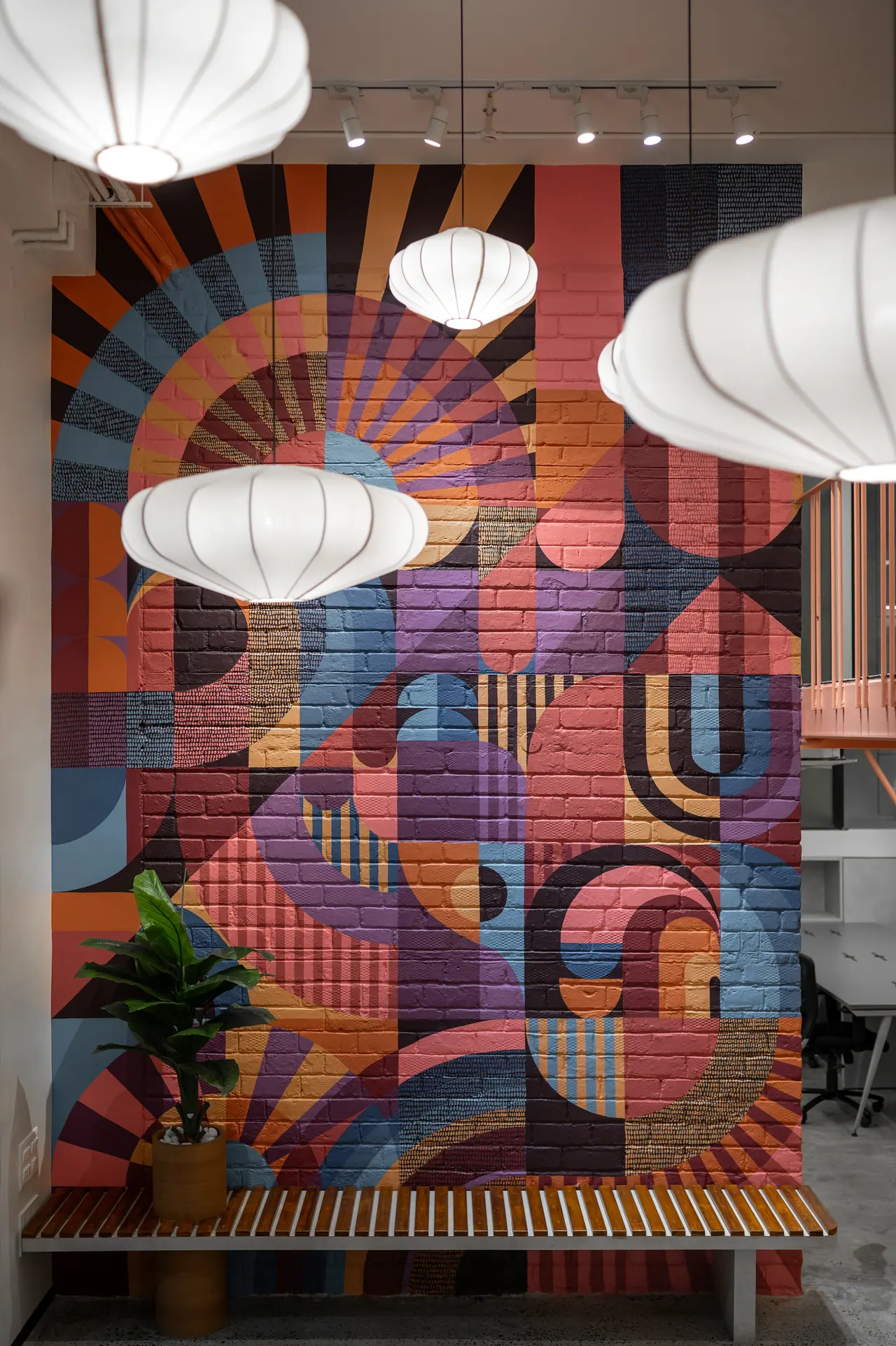 Painted brick mural of orange, pink, purple and blue geometric shapes behind white lantern pendants and a slatted bench