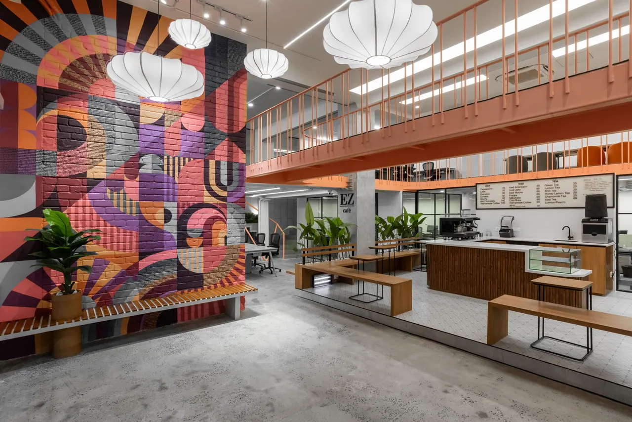 Double-height café with a painted brick mural, white lantern pendants, timber benches and an orange-railed mezzanine