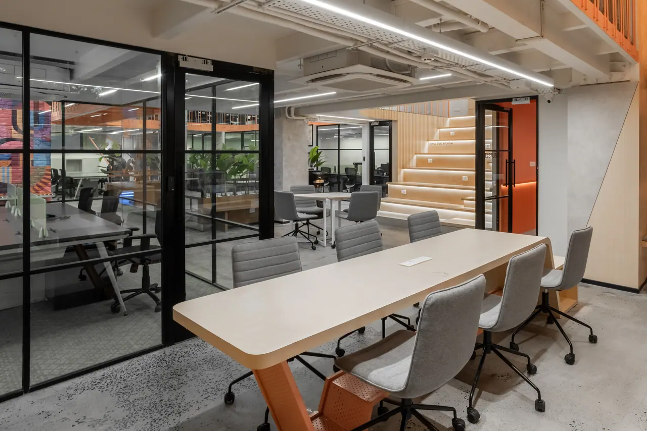 Long pale timber table on an orange perforated base with grey chairs, beside glass-walled cabins and lit steps