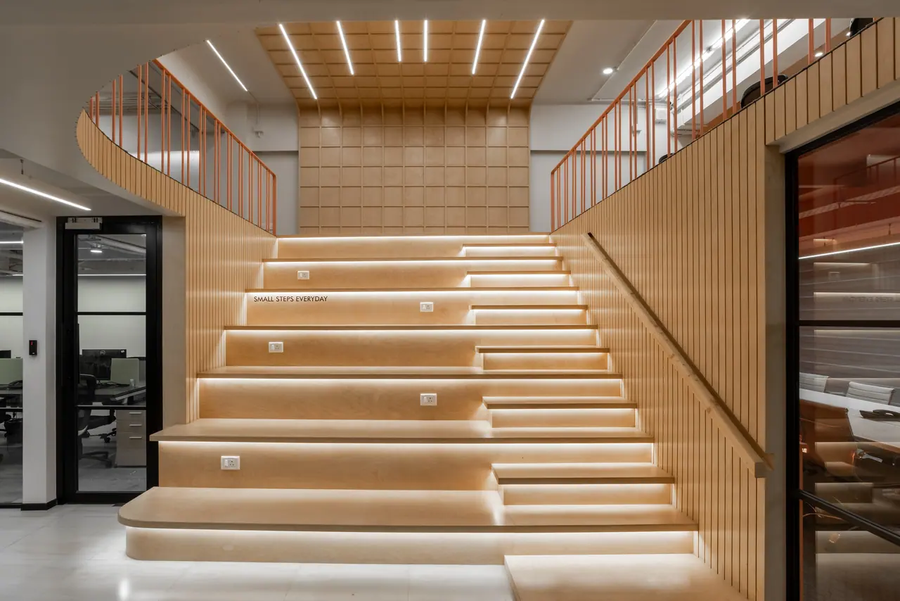 Tiered plywood amphitheatre steps with lit risers and sockets under a coffered timber ceiling and orange railings