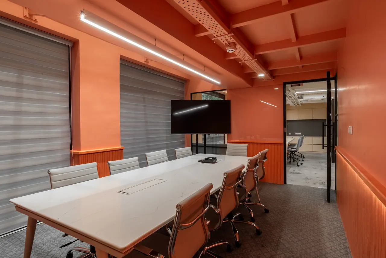 Orange-painted meeting room with a white marble-look table, white leather chairs, a screen on a stand and grey blinds