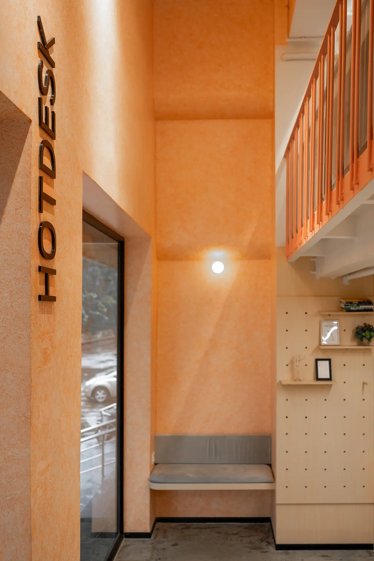 Hot-desk alcove with orange textured walls, HOTDESK lettering, a cushioned bench, pegboard shelves and a tall window