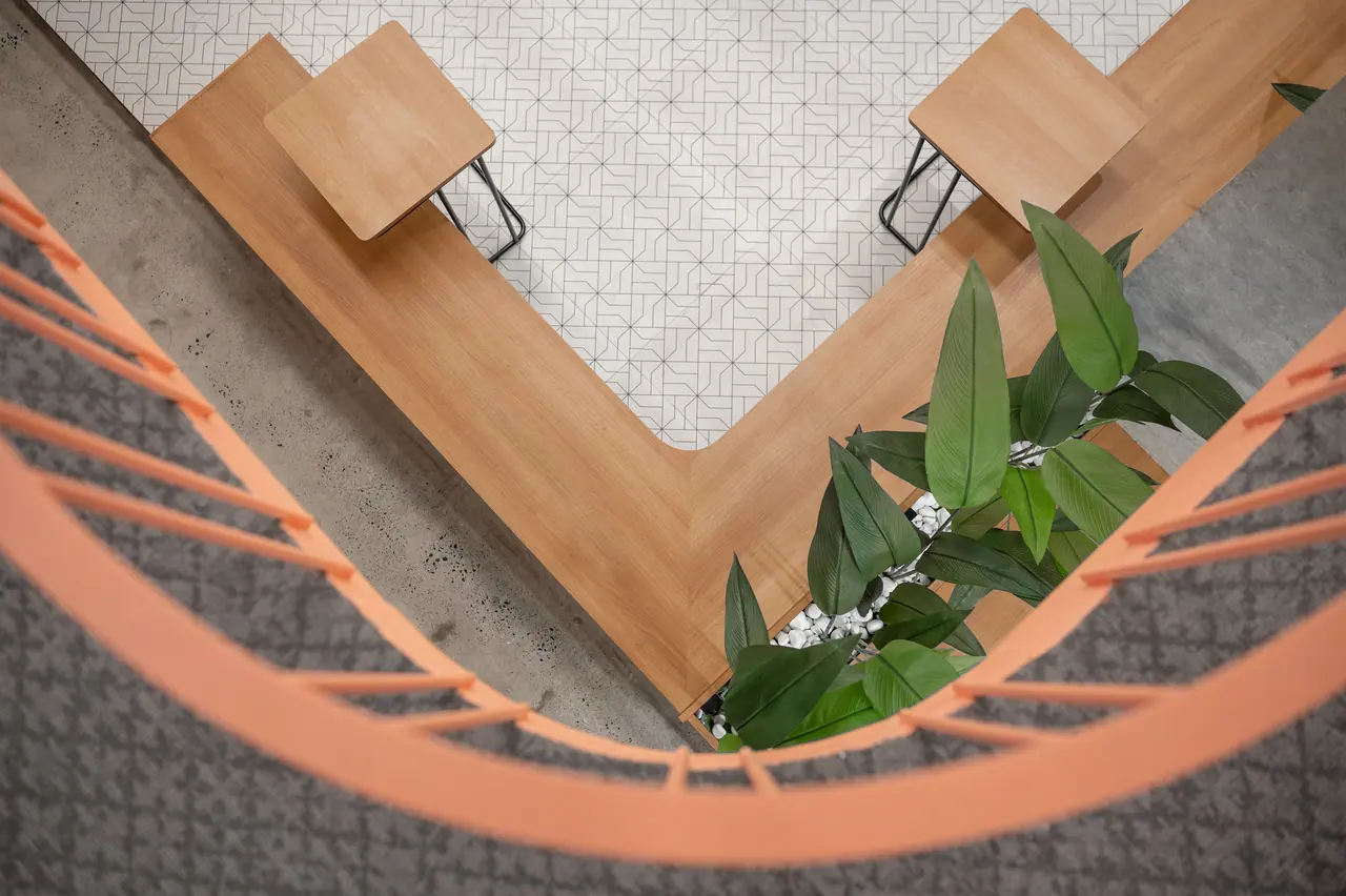 Overhead view through a curved orange railing of a V-shaped timber bench, small tables and plants on patterned tiles