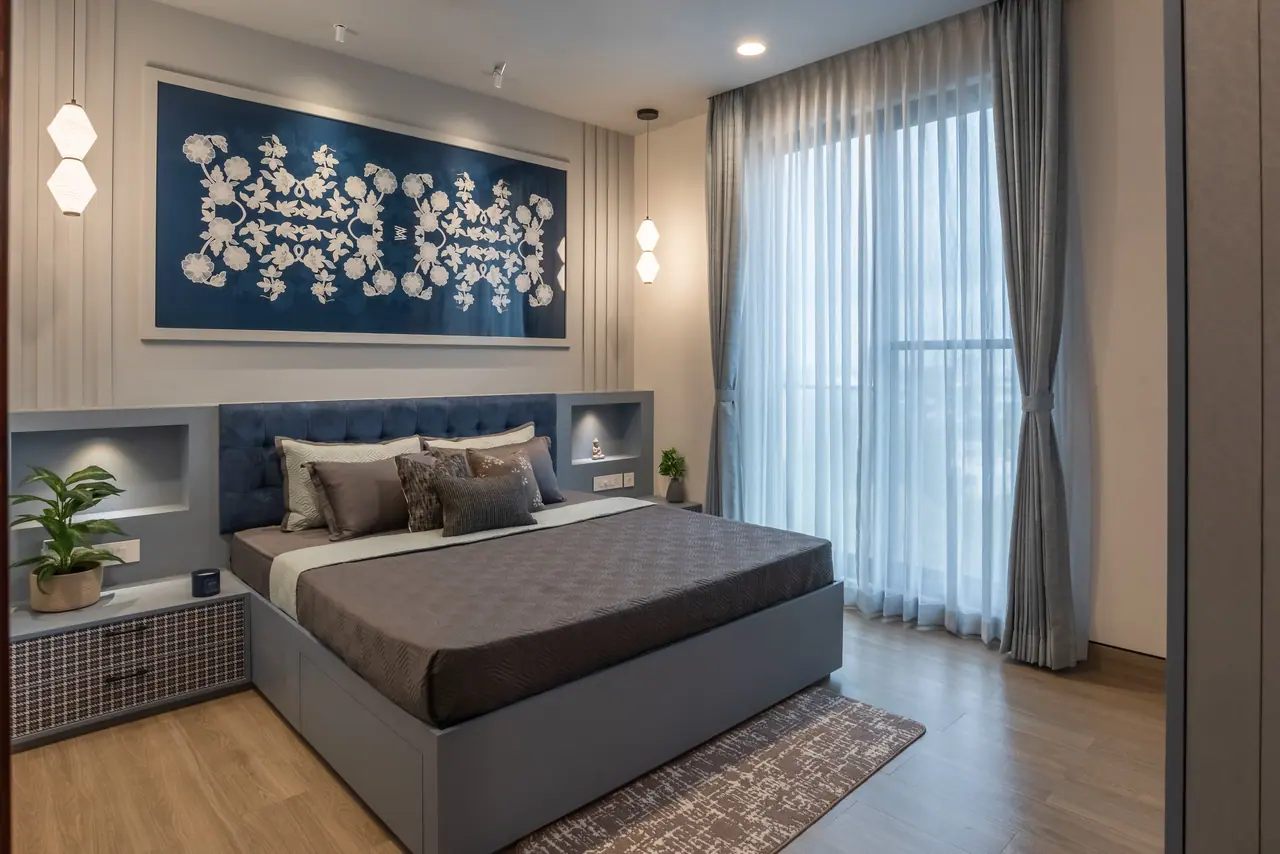 Bedroom with a tufted blue headboard, built-in lit niches, a large indigo floral artwork, white pendants and sheer curtains