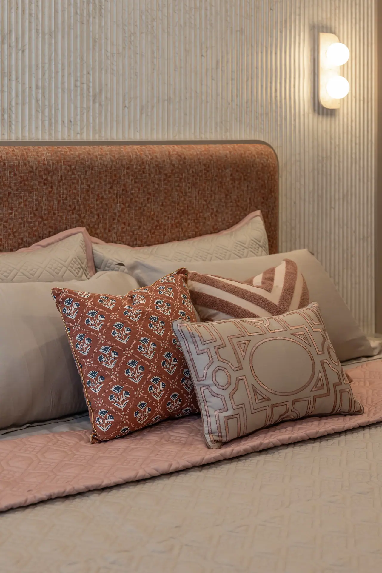 Rust textured headboard with block-print, chevron and geometric cushions on a pink quilt against a fluted wall panel