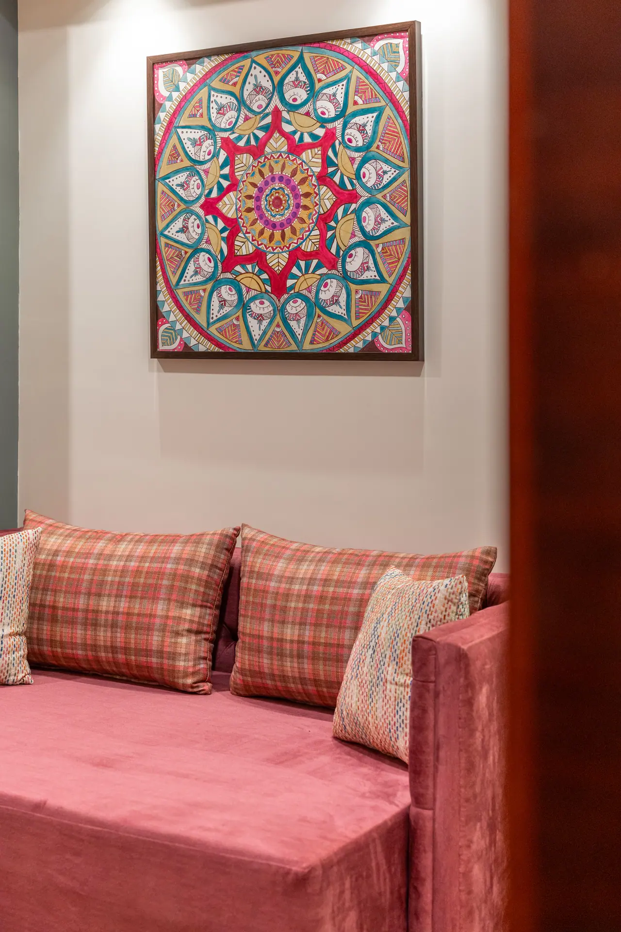 Pink velvet daybed with checked and woven cushions beneath a framed mandala painting in red, teal and gold
