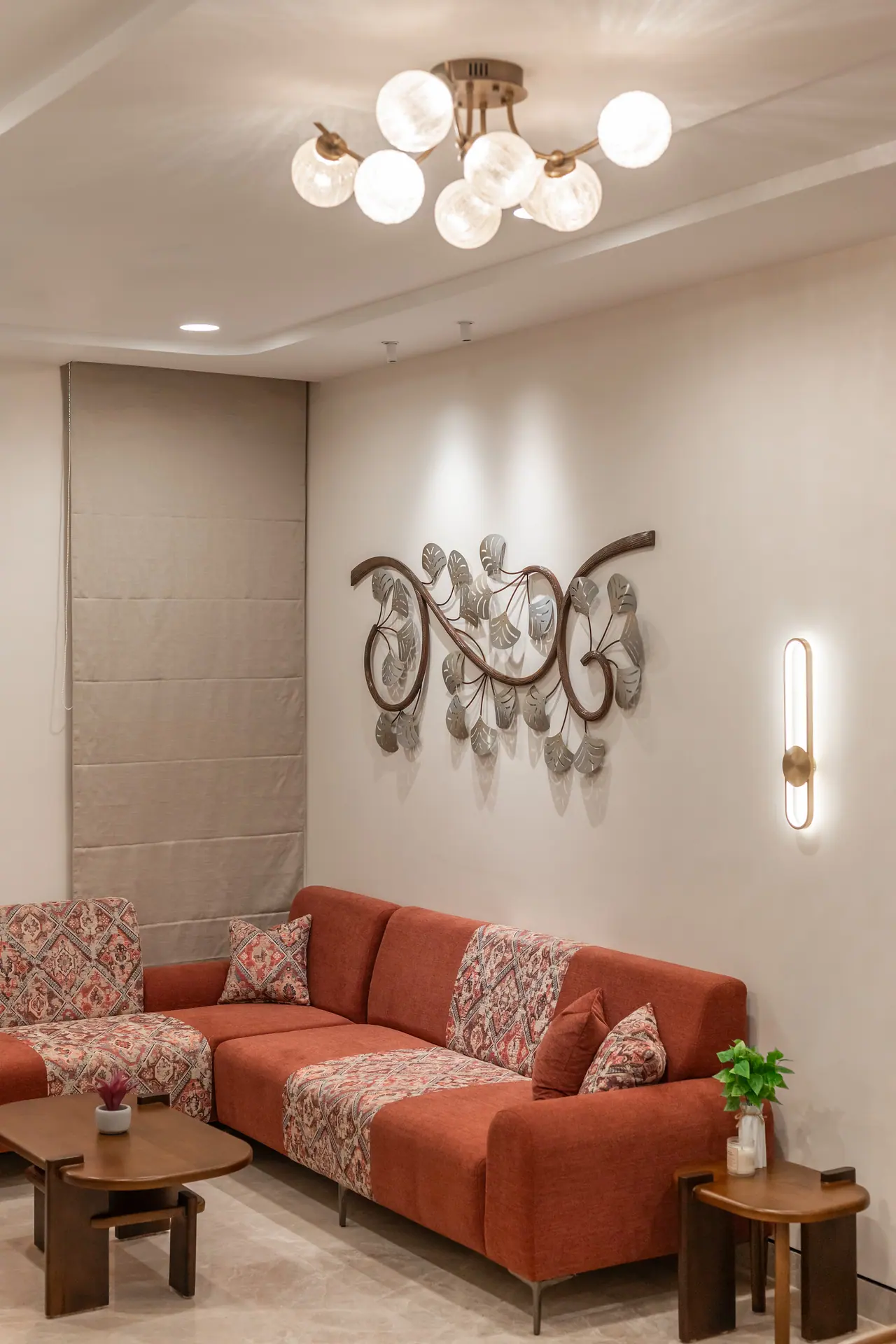 Rust sofa with patterned throws below a glass-globe ceiling light, metal leaf wall art and a slim brass wall light