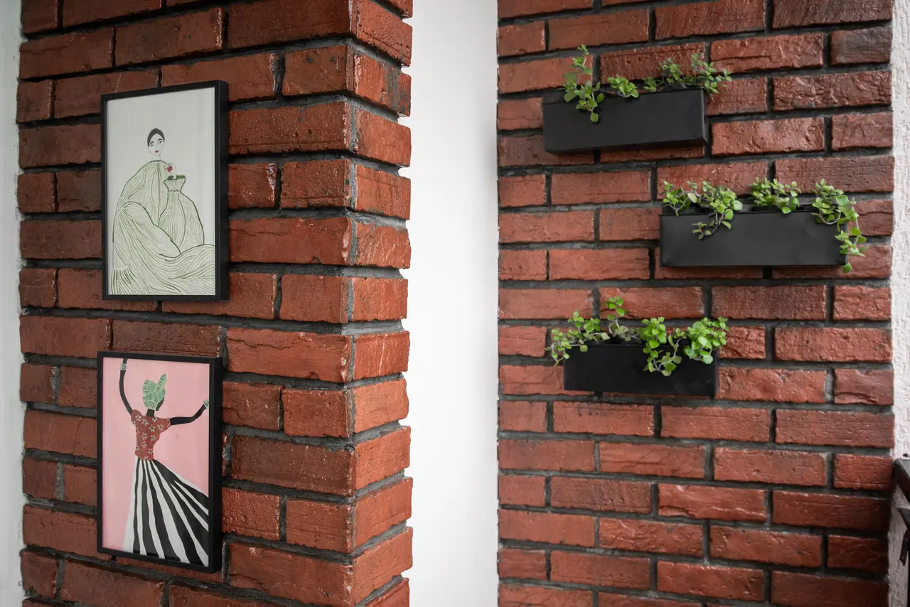 Red brick wall with two framed illustrations of women beside three black wall planters holding trailing green plants