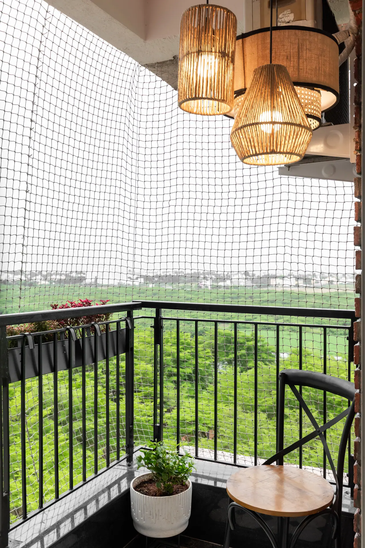 Balcony corner with a bistro chair, potted plant and rattan pendants behind a black railing and net, overlooking greenery