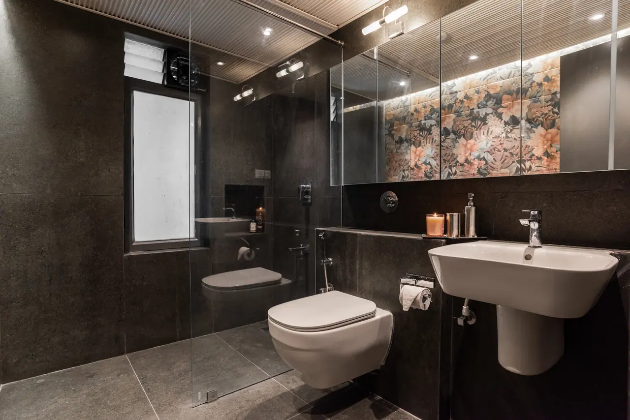 Dark stone-clad bathroom with a glass shower screen, wall-hung WC and basin, and floral tiles lit behind a mirror cabinet