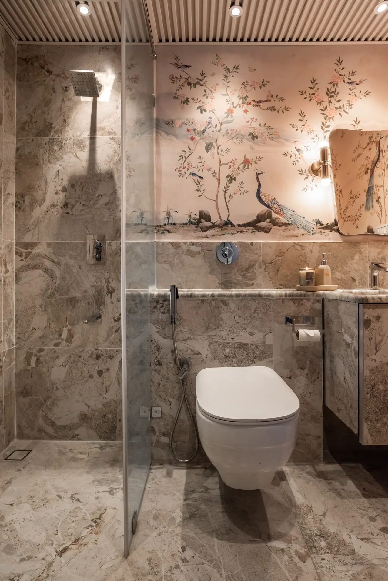 Walk-in shower and wall-hung WC in grey marble, with a peacock and blossom wall print and a brass sconce above the counter