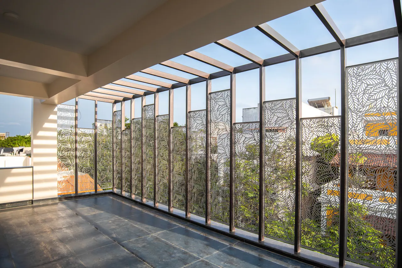 Covered terrace with laser-cut leaf-pattern metal screens under a steel pergola, dark stone floor and greenery beyond