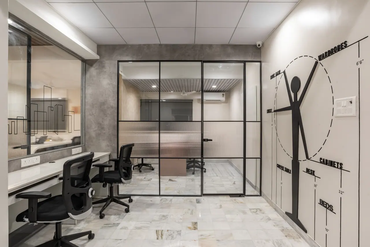 Workstations facing a black-framed fluted glass cabin, with a stick-figure furniture dimension graphic on the right wall