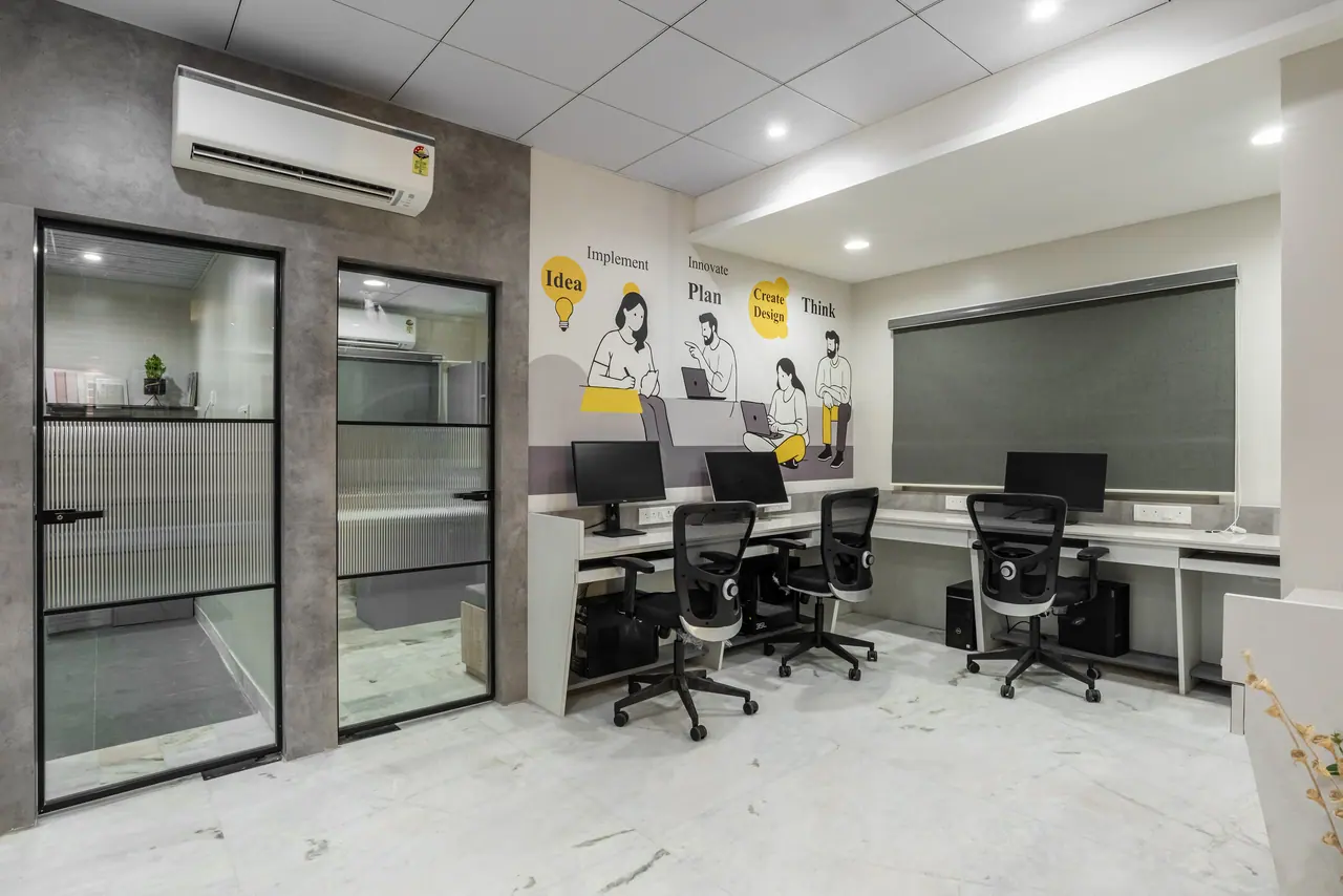 Open workstations with monitors and mesh chairs below an illustrated mural of people at work, beside black-framed glass doors