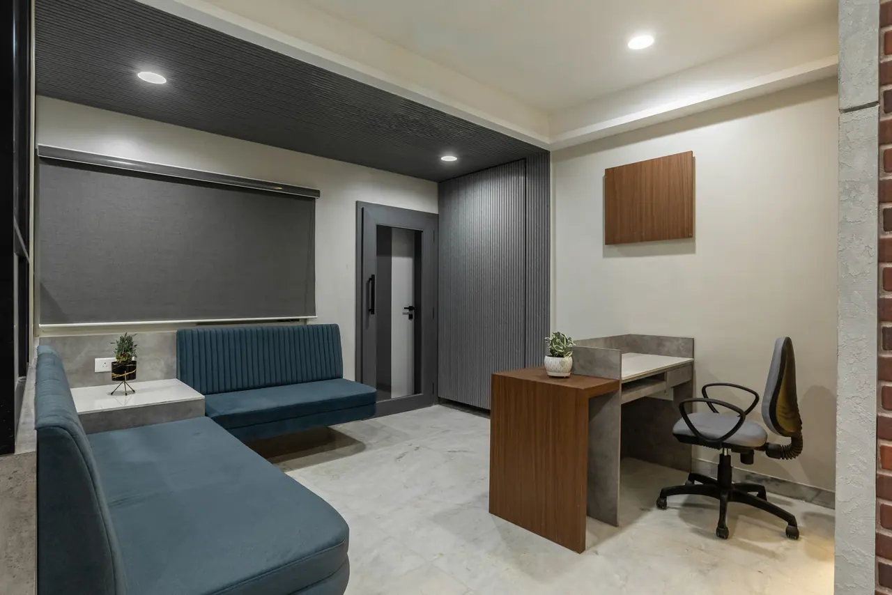 Waiting lounge with an L-shaped teal banquette, side table, roller blind, fluted grey panel and a wood-and-concrete desk