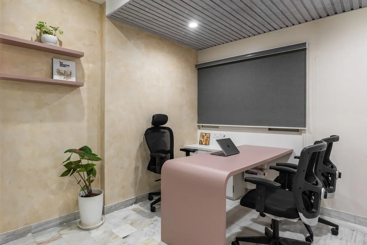 Small cabin with a blush pink waterfall desk, black mesh chairs, limewash walls, floating shelves and a grey slatted ceiling