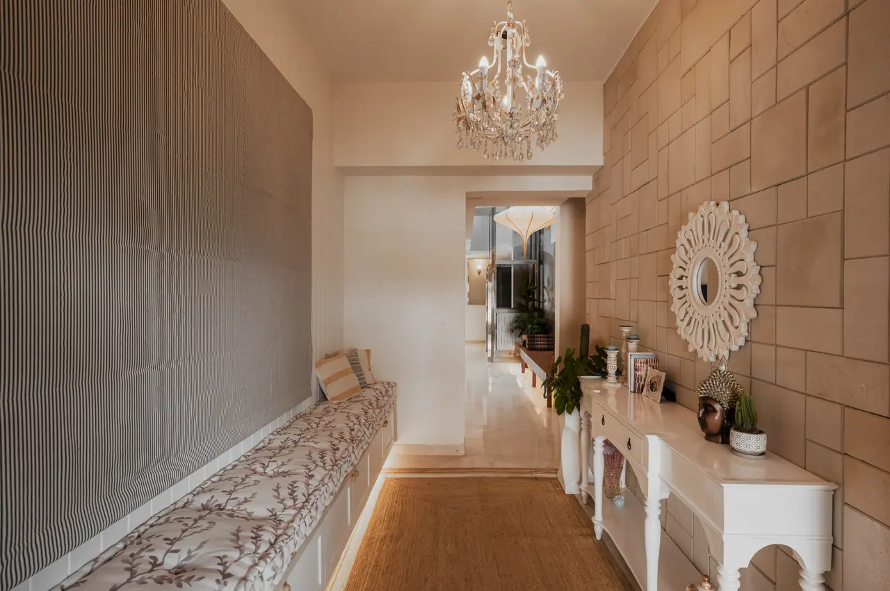 Corridor with a crystal chandelier, cushioned bench seat, white console table, carved round mirror and stone-block wall