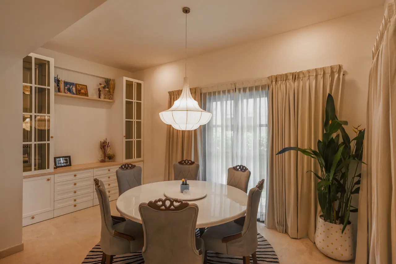 Dining room with a round white table, grey chairs with carved wooden crests, fabric pendant, striped rug and glazed cabinets