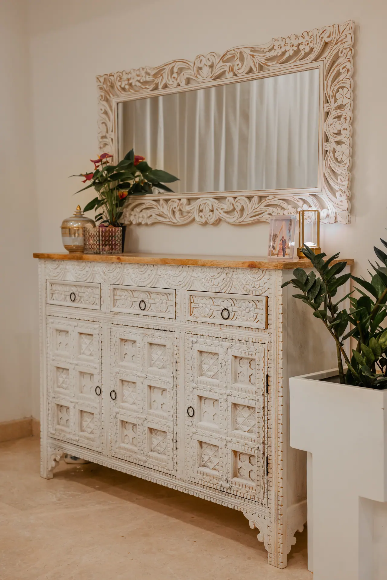 Carved white-painted sideboard with a wooden top, below an ornate carved mirror, with an anthurium and a potted plant